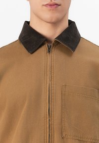 Ungdom som har på seg en brun zip-up jakke med mørkebrun cord-fløyelskrage og brystlomme, vist fra halsen til midten av brystet.