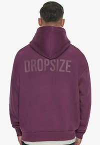 Violetinė džemperis su dideliu, šviesiai pilku "DROPSIZE" spaudu nugaros pusėje. Pagamintas iš minkštos medžiagos, turi laisvą siluetą ir reguliuojamą kapušoną.