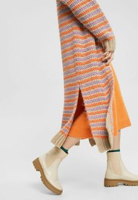 Stickad cardigan med lila, orange och beige ränder; orange kjol; beige ankellånga stövlar med kraftiga sulor och gröna accenter. Sidoflikar synliga.