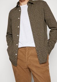 Camicia button-up marrone con un motivo, con una texture sottile, indossata su una maglietta bianca, abbinata a pantaloni in corduroy beige.