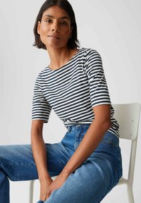 Femme aux cheveux courts et foncés portant une chemise à rayures noire et blanche et un jean bleu, assise sur une chaise blanche contre un fond uni.