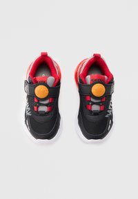 Ténis pretos e vermelhos com materiais em rede e sintéticos, design curvo, tira de velcro, detalhes cinza e um pormenor circular amarelo.