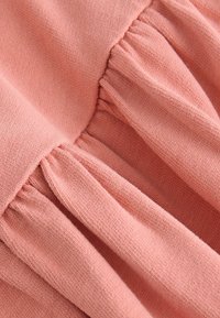 Helles Rosa, strukturiertes Gewebe, das Falten und Rüschen zeigt und einen sanften, drapierten Effekt erzeugt. Das Material scheint gestrickt mit einer leicht groben Webart.
