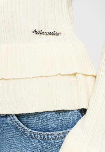 Pulover crem împăturit cu un tiv volan și un accent cu logo argintiu pe față, asociat cu blugi din denim de culoare albastru deschis.