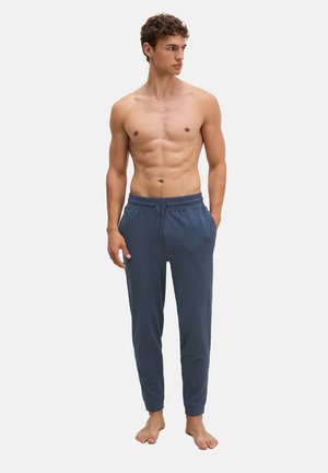 Oberkörperfreier junger Mann mit muskulösem Körperbau, trägt blaue Jogginghosen mit Kordelzug, steht barfuß vor einem schlichten hellen Hintergrund.