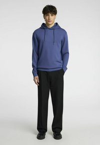 Felpa con cappuccio blu dotata di lacci regolabili, abbinata a pantaloni neri con una vestibilità morbida e una texture liscia. Le scarpe nere completano l'outfit.