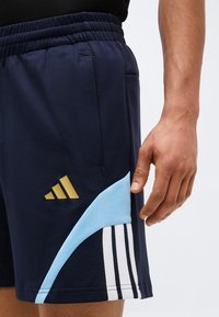 Κοντινό πλάνο του χεριού ενός ατόμου δίπλα σε σκούρο μπλε αθλητικό σορτς με ανοιχτό μπλε και λευκές ρίγες και ένα χρυσό λογότυπο στον μηρό.