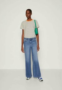 Woman wearing striped T-shirt with orange neckline, wide-leg blue jeans, white sneakers, and a green shoulder bag, standing against plain background.