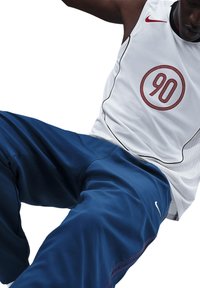 Witte sportieve tanktop met een rode cirkelvormige "90" graphic en zwarte bies, gecombineerd met donkerblauwe broeken met een wit Nike-logo.