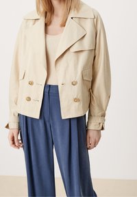 Beżowa krótka kurtka o podwójnym zapięciu, z dużym kołnierzem, przednimi kieszeniami i złotymi guzikami, w zestawie z niebieskimi spodami z plisami.