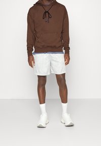 Felpa marrone con cappuccio e cordini, tasca a marsupio, indossata sopra una maglietta blu, abbinata a pantaloni corti grigio chiaro e sneakers bianche chunky.
