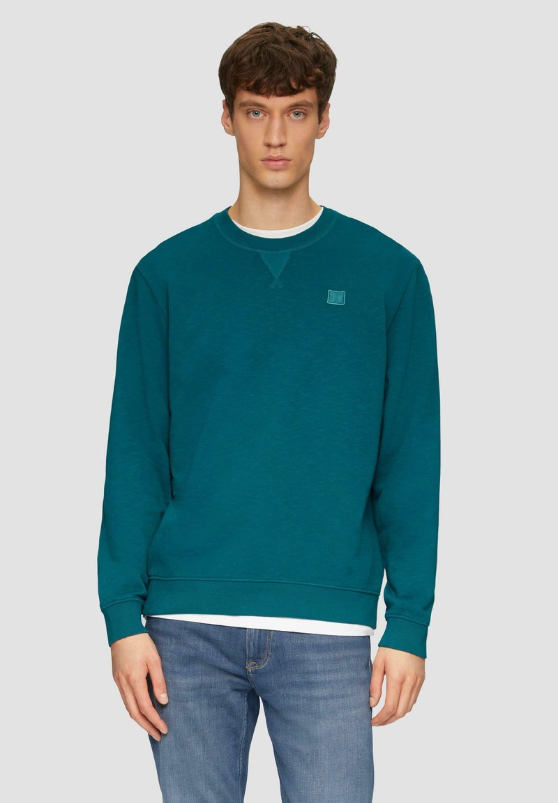Felpa crewneck color turchese realizzata in morbido tessuto, con un piccolo patch del logo sul petto, polsini e orlo a coste, indossata sopra una camicia bianca.