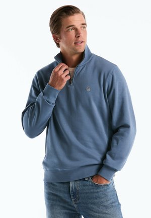 Homme portant un sweat-shirt bleu à fermeture éclair avec un logo de pingouin, main près de la fermeture éclair, regardant vers la droite, vêtu d'un jean bleu clair sur fond blanc.