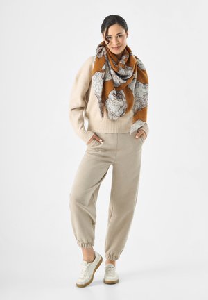 Beige trui met geribbelde zoom, crèmekleurige joggingbroek met elastische manchetten, en een oversized sjaal in een gemengd patroon van oranje en grijs. Witte sneakers maken de look compleet.