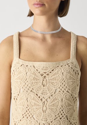 Beżowy top na ramiączkach z szydełka z motywem motyli i grubymi ramiączkami, w zestawieniu ze srebrnym metalicznym chokarem na szyi.