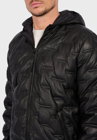 Homme portant une veste en cuir matelassé noir avec des coutures horizontales, fermeture éclair à l'avant, poches zippées et capuche attachée sur une chemise blanche.