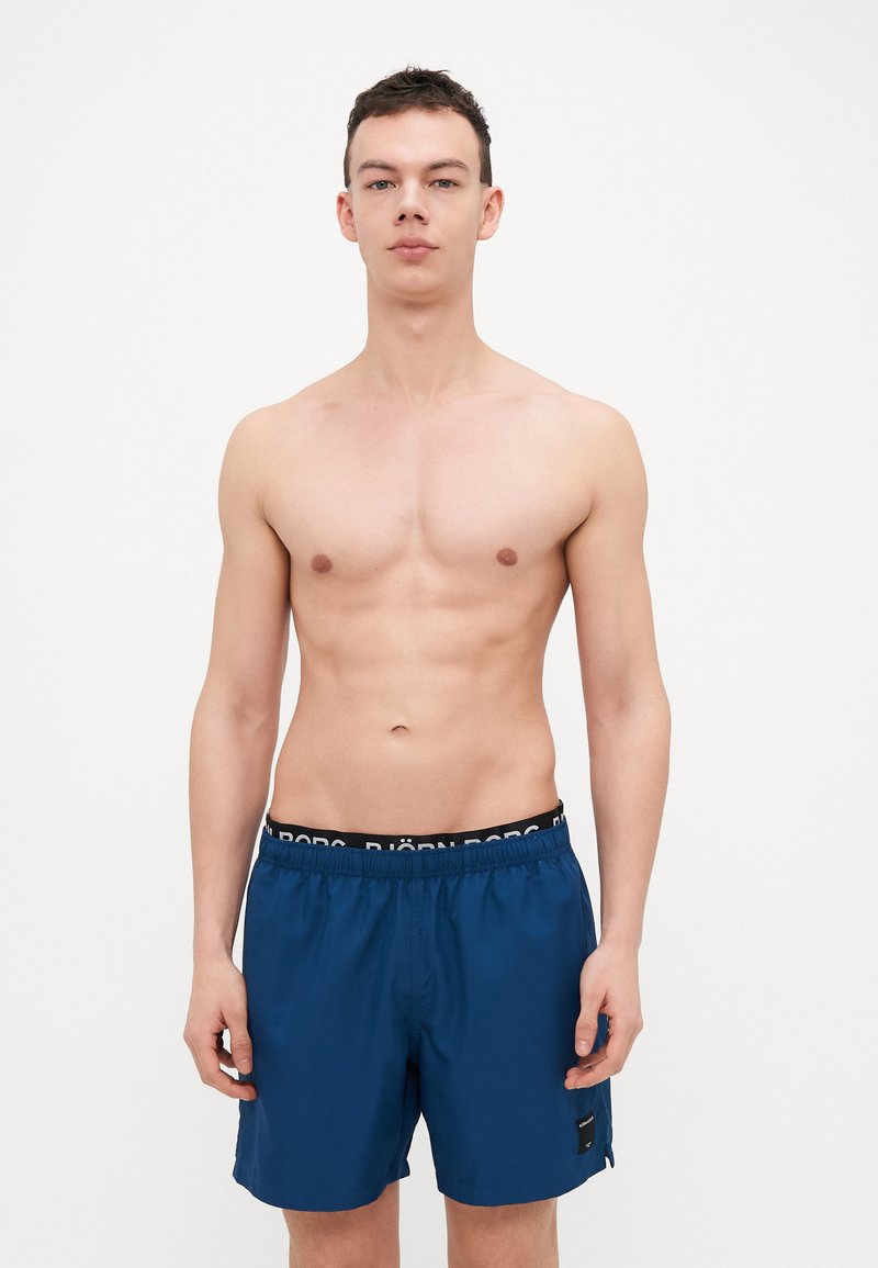Blue swim shorts made from lightweight fabric with an elastic waistband featuring a black logo. Straight cut and above-knee length.