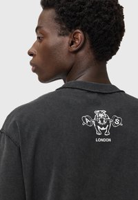 Camiseta gris de gran tamaño con mangas cortas. Presenta un gráfico de un perro musculoso sosteniendo pesas en la parte posterior, y el texto "LONDRES" debajo.