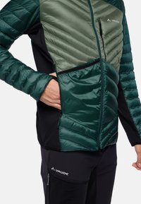 Padded Jacke in Dunkelgrün und Hellgrün, mit einer Reißverschlusstasche, vertikalen Nähten und schwarzen Stoffakzenten an Ärmeln und Torso.