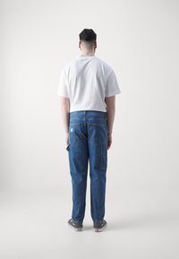 Camiseta de algodón blanca y pantalones cargo de mezclilla azules con bolsillos, usados con zapatillas grises. El modelo está de espaldas a la cámara.