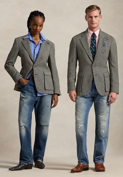 Deux mannequins posent debout, vêtus de blazers gris à chevrons, de jeans bleus et de chaussures habillées ; l’un avec une chemise bleue, l’autre avec une chemise et une cravate.