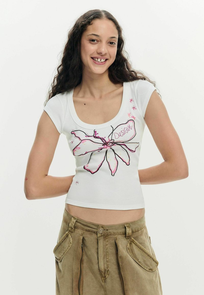 Jeune femme aux cheveux longs et bouclés souriante, portant un crop top blanc avec un motif floral rose et un pantalon cargo beige.
