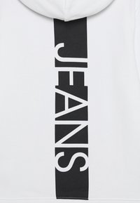 Vit hoodie med en stor vertikal svart "JEANS"-tryck på mitten av ryggen. Mjukt tyg med en strukturerad yta och en dragsko i huvan.