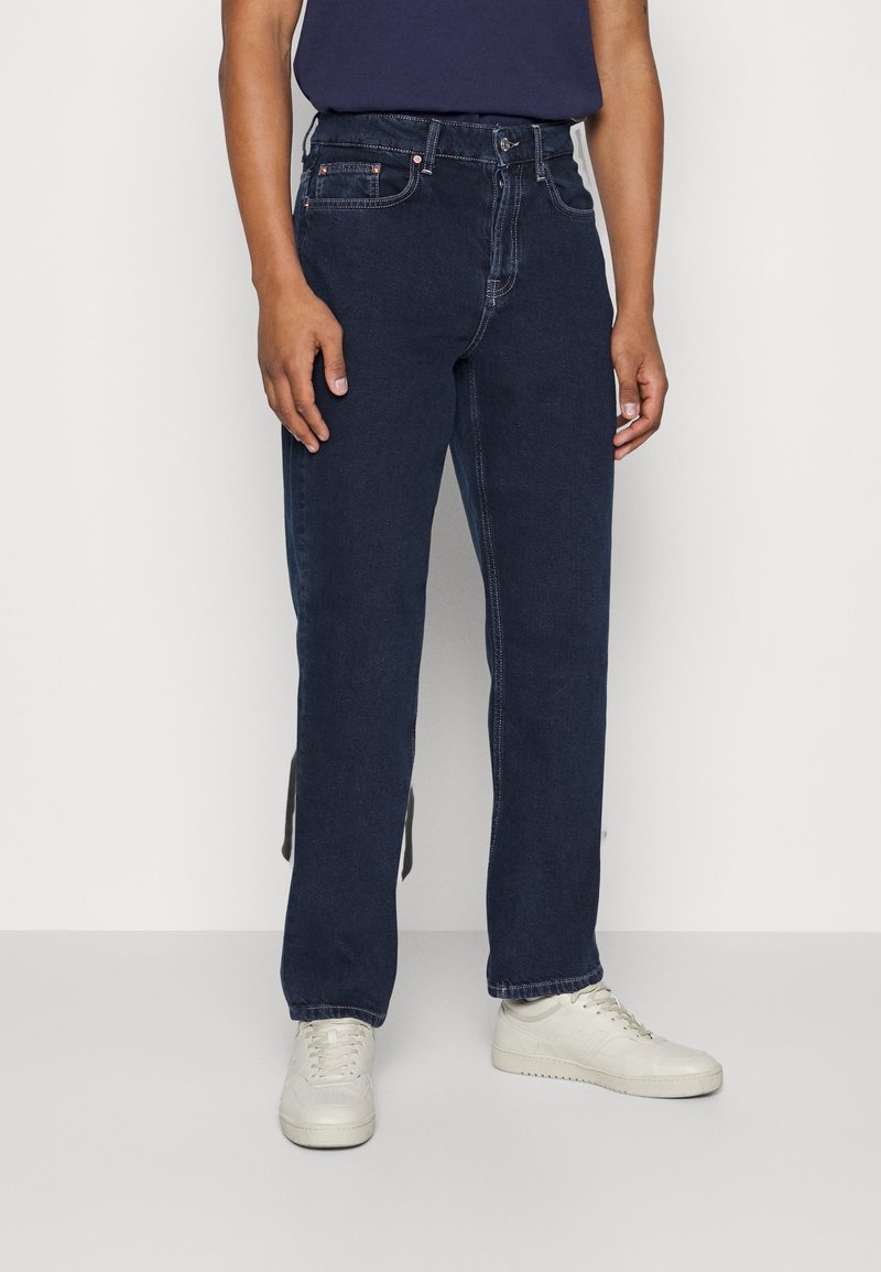 Mörkblå denimjeans med rak bendesign, med orange sömmar, fem fickor och knappgylf. Vita sneakers under.