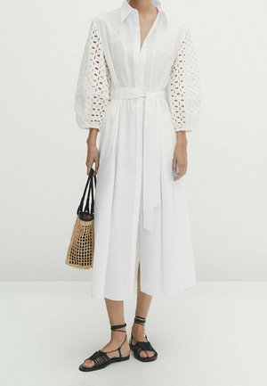 Robe blanche avec des manches ballon volumineuses et brodées, taille cintrée, longueur mi-mollet, assortie à un sac en osier et des sandales noires à lanières.