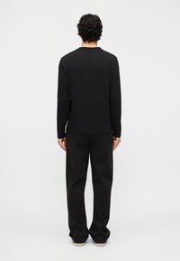 Άνδρας με σγουρά μαλλιά στέκει γυρισμένος με την πλάτη, φορώντας μαύρο μακρυμάνικο πουκάμισο, μαύρο παντελόνι και λευκά παπούτσια πάνω σε απλό φωτεινό φόντο.