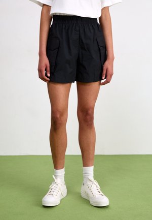 Personne portant un short noir avec des poches cargo, des chaussettes blanches et des baskets blanches, debout sur un sol vert contre un mur blanc.