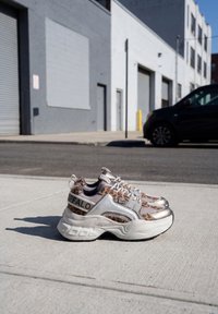 Χοντρά λευκά και λεοπάρ sneakers με μεταλλικές λεπτομέρειες που ακουμπούν σε έναν ηλιόλουστο τσιμεντένιο πεζοδρόμιο δίπλα σε δρόμο με παρκαρισμένο αυτοκίνητο.