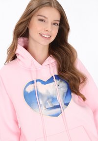 Rózsaszín kapucnis pulóver, amelyen egy nagy szív alakú grafika látható, fehér állattal kék háttér előtt; puha anyagból készült, húzózsinórokkal.