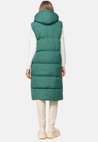 Langer, kapuzenloser Puffer-Westen in Türkis mit horizontalen Steppungen. Gestylt mit einem weißen Langarmshirt und cremefarbenen, platten, klobigen Stiefeln.