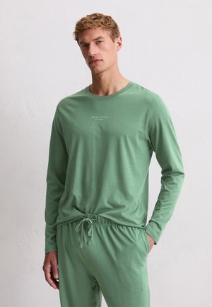 Junger Mann in einem salbeigrünen Langarmshirt und passenden Jogginghosen, der mit einer Hand in der Tasche vor einer schlichten Wand steht.