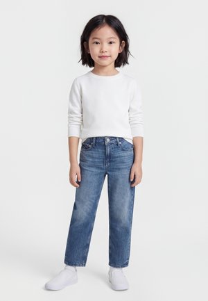 Bambino con capelli corti neri, indossa una maglia bianca a maniche lunghe, jeans blu e sneakers bianche, in piedi davanti a uno sfondo semplice.