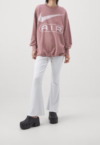 Př oversized růžové mikině Nike s bílým logem, stažením na spodním okraji; v kombinaci s bílými ribbed zvonovými kalhotami a černými dřeváky s otvory.