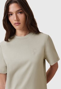 T-shirt beige chiaro in cotone morbido con scollatura rotonda, maniche corte e un piccolo logo ricamato sul petto. Texture liscia.