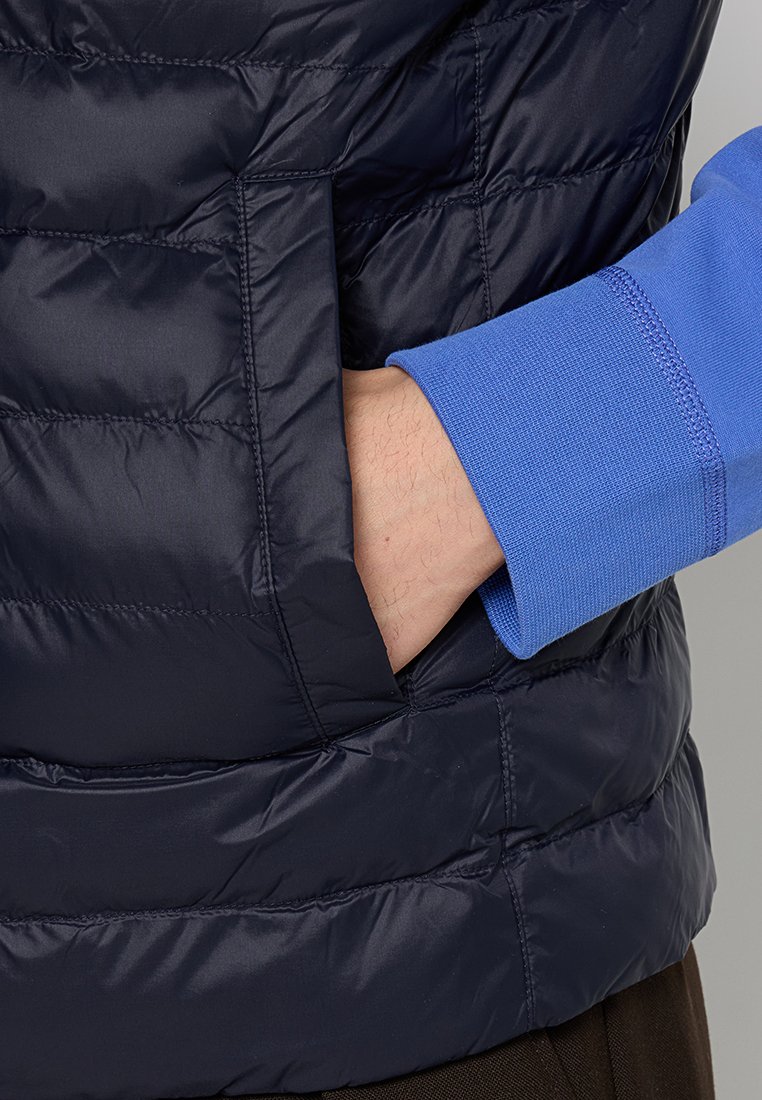 Ręka w bocznej kieszeni granatowej pikowanej kurtki, pod którą ma na sobie długą niebieską koszulę.