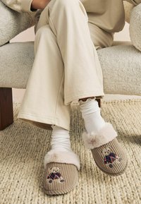 Tan corduroy slippers with a plush, light beige fur cuff. Features a teddy bear embroidered in colorful argyle clothing.