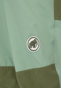 Hellgrünes und olivgrünes Stoffpaneel mit einem kreisförmigen, bestickten Mammut-Logo-Patch, das auf dem helleren Stoffabschnitt aufgenäht ist.