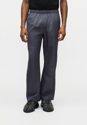 Uomo che indossa pantaloni grigi scuro, larghi, con vita elasticizzata e scarpe sportive nere, in piedi davanti a uno sfondo semplice.