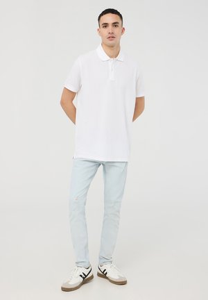Polo bianco a maniche corte, colletto a coste e patta con due bottoni, abbinato a jeans azzurri strappati e sneakers bianche.