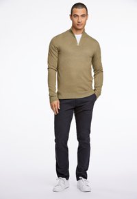 Helles olivgrünes Strickpullover mit einem Reißverschlusskragen, kombiniert mit dunkelblauen Hosen und weißer Sneakers, die einen lässigen Sitz und Textur zeigen.