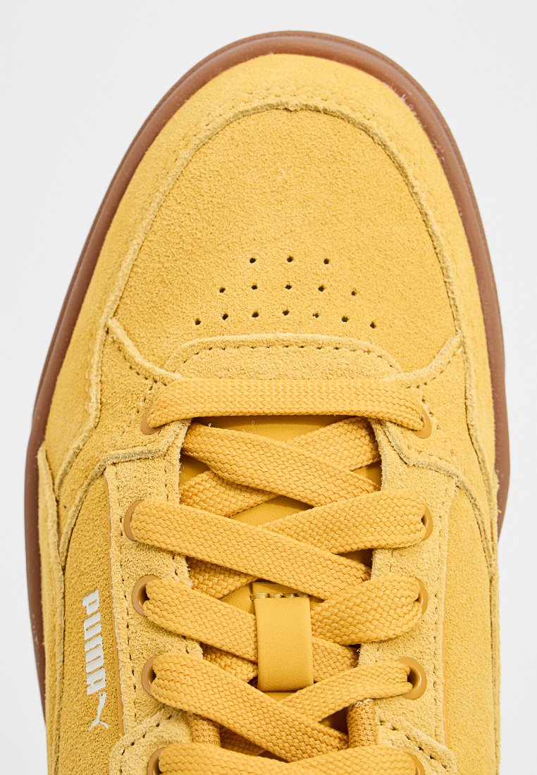 Zapatilla de ante amarilla con suela de goma. Presenta un diseño con cordones, caja de dedos perforada y logo en relieve. Material texturizado en toda la superficie.