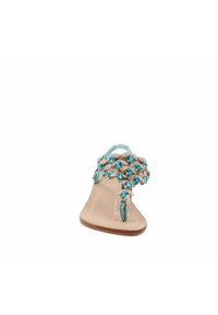 Sandalo piatto con cinturini in beige e gemme finte turchesi sulla parte superiore decorativa. Cinghie trasparenti e una suola piatta completano il design.