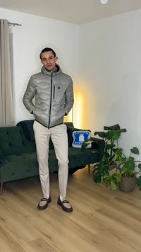Homme portant une doudoune argentée, un pantalon beige et des mocassins bicolores, debout à l’intérieur près d’un canapé vert avec des plantes et un coussin.