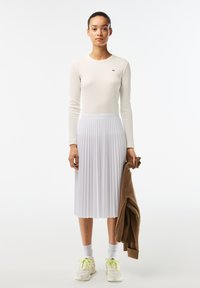 White ribbed long-sleeve top, pleated white midi skirt, brown sweater in hand, white sneakers with neon accents, light gray socks.