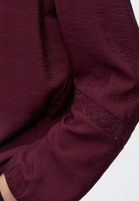 Manche d’un vêtement bordeaux avec tissu texturé et une bande de dentelle près du coude, finition avec un poignet élastique.