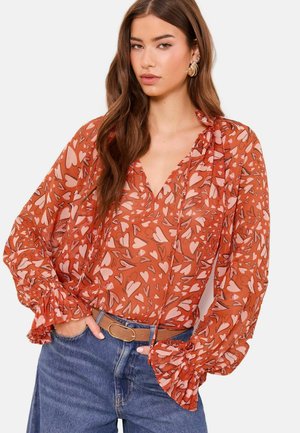 Pitkätukkainen nainen, jolla on punainen paita vaaleanpunaisilla sydänkuvioilla, röyhelöityillä kalvosimilla, puettu sinisiin farkkuihin ja ruskea vyö.